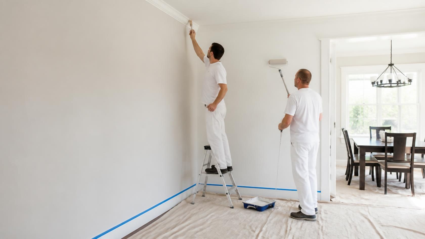Two painters cutting in crown molding and rolling walls in a dining room protected with drop cloths